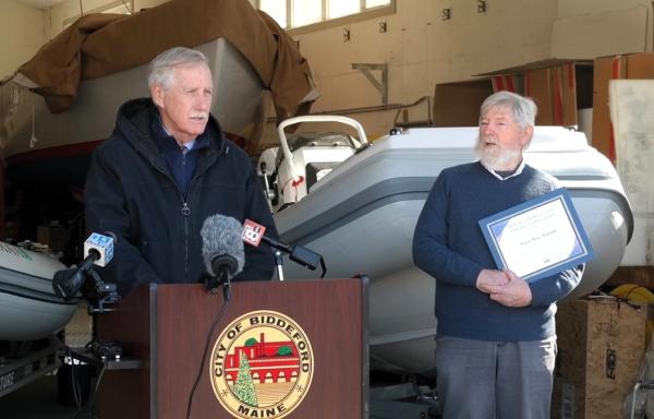 Senator Angus King stands at a podium bearing the seal of the City of Biddeford. Sean Tarpey stands to the right in the photo, holding an award signifying Saco Bay Marine's REAP grant. In the background are 2 electric boats and a larger boat in the far background.