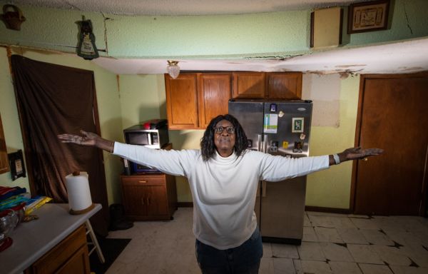 A women standing in her house with her arms held out at shoulder height to show how dipped her roof is. It sags in the middle where she stands.