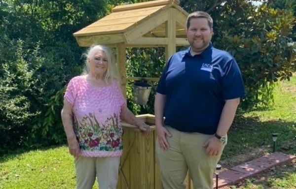 Smiling elderly woman next to a younger man standing in front of a wooden structure that covers a well in a green lawn.