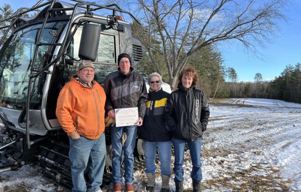 Two men and two women stand in front of a snow grooming machine in a snowy field under a blue sky. They are dressed warmly, and there are leafless trees in the background.