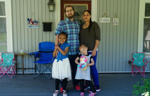 A smiling family, father, mother, two young daughters, standing on the porch of their own home.