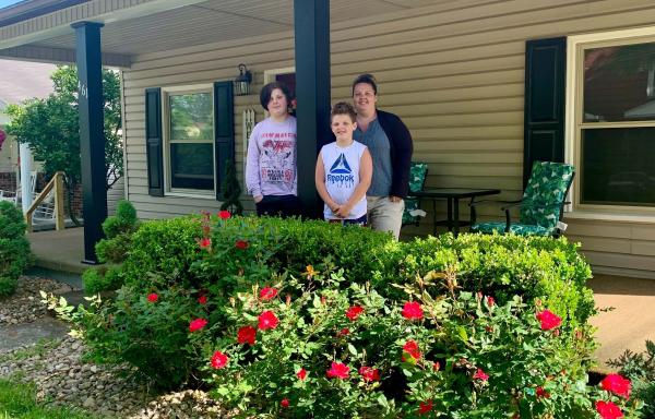 Mom and two sons standing on the porch of their two-story house, as seen from the yard,