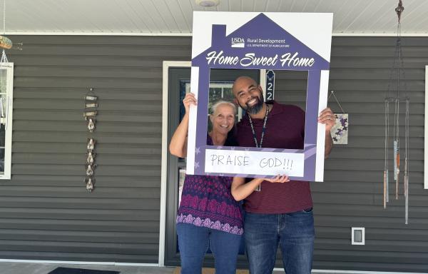 Ariel and Mike holding a home sweet home picture frame in front of Ariel's home
