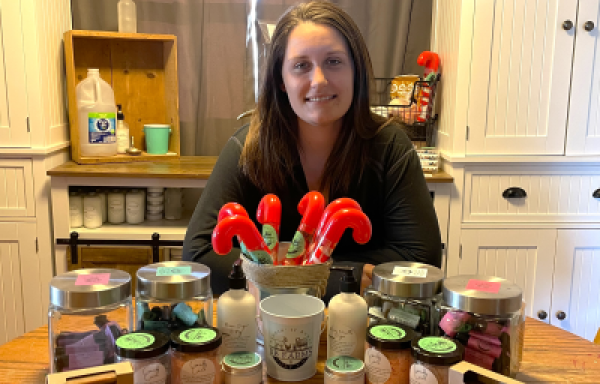 Woman sitting at table with home made soaps, lotions, and balms.