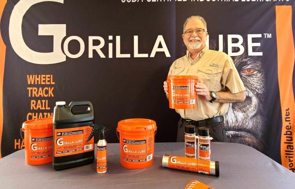 A bearded man poses in front of a backdrop that says "GORiLLA LUBE" with industrial products on a table in front of him with orange labels. 