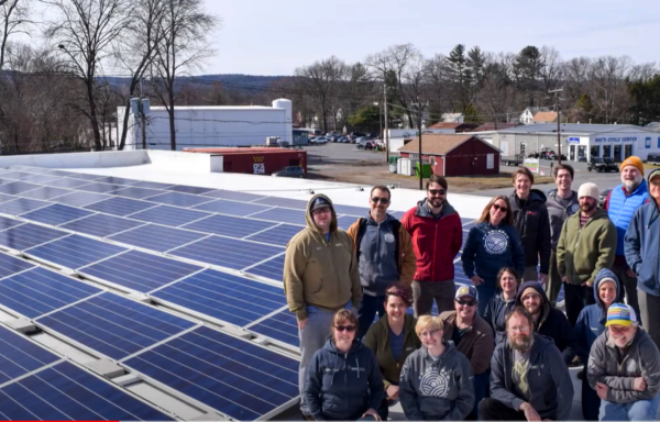 Members of the Real Pickle Co-op stand on the roof of the building next to the new solar panels on a sunny day. Many are wearing coats and it looks chilly outside.