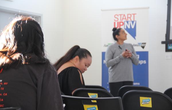 picture of presenter looking off frame towards a projector screen. behind the presenter is a banner that says startup nv.